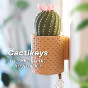Puede incluir: Un porta llaves beige con forma de maceta de cactus con un cactus verde y una flor rosa en la parte superior. El porta llaves está montado en una pared y tiene una llave unida a él. El texto "Cactikeys The Blooming Keyholder" es visible debajo del porta llaves.