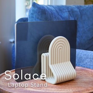 Può includere: Un supporto per laptop color crema con un design ad arco. Il supporto è composto da linee curve e la parola "Solace" è stampata sulla parte anteriore. Il supporto è su un tavolo di legno, con un laptop dietro.