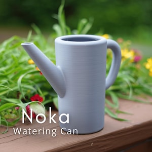 Puede incluir: Una regadera gris claro con un asa y un pico curvos. El texto "Noka Watering Can" se muestra en blanco. La regadera está sobre una superficie de madera marrón con follaje verde y flores coloridas en el fondo.