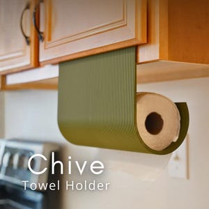 Puede incluir: Portarrollos de papel de cocina verde oliva montado debajo de un armario de madera. El soporte tiene un diseño curvo con estrías verticales y el texto "Chive Towel Holder" en blanco. Se ve un rollo de papel de cocina.