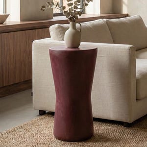 Könnte beinhalten: Ein weinroter Beistelltisch mit einer Vase mit getrockneten Zweigen. Der Tisch hat eine geschwungene Form. Die Vase ist beige und hat einen kleinen Griff. Der Tisch steht auf einem gewebten Jute-Teppich neben einem beigen Sofa.
