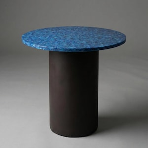Op de afbeelding: Een moderne bijzettafel met een rond blad en een cilindrische voet. Het tafelblad is een mozaïek van blauwtinten, terwijl de voet donkerbruin is. De tafel heeft een diameter van ongeveer 50 cm en is 61 cm hoog.
