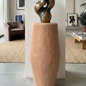 Könnte beinhalten: Eine abstrakte Bronzeskulptur mit fließender Form steht auf einem hohen, terrakottafarbenen Sockel. Die Skulptur hat eine polierte, reflektierende Oberfläche. Der Sockel ist zylindrisch und strukturiert und verleiht dem modernen Kunstwerk ein rustikales Element.