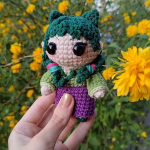 Puede incluir: Una muñeca de crochet con pelo verde, un vestido verde y morado y grandes ojos negros. La muñeca está siendo sostenida en una mano contra un fondo de flores amarillas.