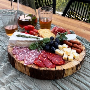 May include: A wooden charcuterie board with an assortment of meats, cheeses, crackers, and grapes. The board is surrounded by glasses of amber-colored liquid, a clear pitcher, and fresh greenery. The scene is set on a wooden table with a green cloth.