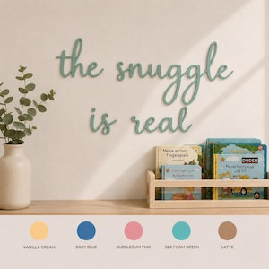 May include: A sea foam green wall sign with the cursive text "the snuggle is real" mounted above a wooden shelf containing children's books. Below the shelf, five color circles are labeled: vanilla cream, baby blue, bubblegum pink, sea foam green, and latte.