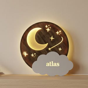 Könnte beinhalten: Nachtlicht aus Holz mit einem Halbmond, Sternen und einer Wolkenform. Der Name "atlas" ist auf der Wolke zu sehen. Das Nachtlicht hat ein warmes Leuchten und ist für ein Kinderzimmer konzipiert.