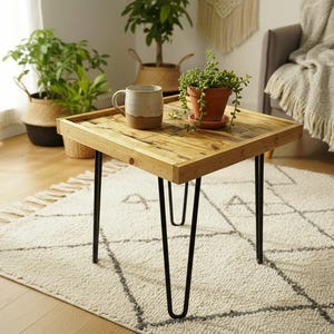 Könnte beinhalten: Ein quadratischer Couchtisch aus Holz mit schwarzen Haarnadelbeinen. Die Tischplatte besteht aus hellem Holz mit sichtbarer Maserung. Eine Keramiktasse und eine Topfpflanze stehen auf dem Tisch. Der Tisch steht auf einem gemusterten Teppich.