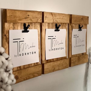 May include: Three wooden display boards mounted on a wall, each with a black clip holding white paper. The paper on each board features the text "Made by HORNTTON" in black. The wood has a natural, light brown finish.