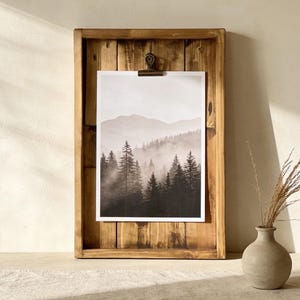 May include: A framed landscape print featuring a black and white forest scene with mountains in the background. The print is held in a rustic, rectangular wooden frame with a metal clip. A small, beige vase with dried plants sits nearby.