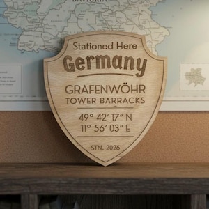 Może przedstawiać: Jasnobrązowa drewniana tablica w kształcie tarczy z napisem "Stationed Here Germany GRAFENWÖHR TOWER BARRACKS" i współrzędnymi. Tablica stoi na ciemnej drewnianej półce, a w tle widoczna jest mapa.