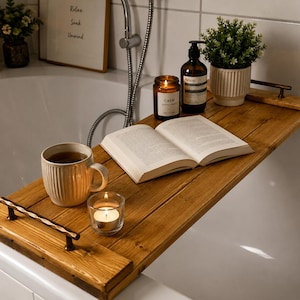 Peut inclure: Un plateau de baignoire rectangulaire en bois doté de deux poignées en métal foncé aux extrémités. Le plateau supporte un livre ouvert, une tasse en céramique blanche, un bougeoir en verre avec une bougie allumée, un flacon en verre et une plante.