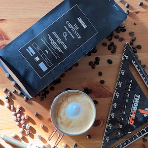 Könnte beinhalten: Draufsicht auf einen schwarzen Kaffeebeutel "The Carpenter" mit Kaffeebohnen, die um eine Kaffeetasse mit einem Milchschaummuster verstreut sind. Ein Metalllineal und ein Schraubendreher sind ebenfalls vorhanden.
