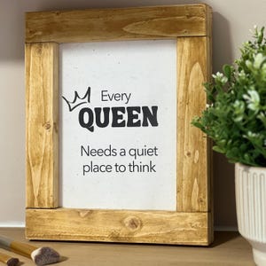 Könnte beinhalten: Ein gerahmtes Holzschild mit dem Text "Every QUEEN Needs a quiet place to think" in schwarzer Schrift. Das Schild hat einen hellbraunen Holzrahmen und einen weißen Hintergrund. Eine kleine Krone befindet sich über dem Wort "QUEEN".