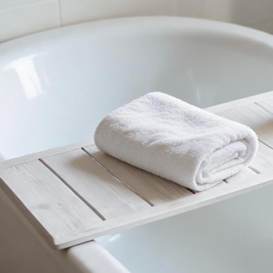 May include: A rolled white towel rests on a white wooden bath tray, positioned over a white bathtub. The tray has a slatted design, and the scene is bathed in soft, natural light, creating a spa-like ambiance.