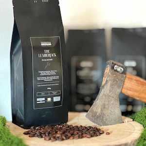 Könnte beinhalten: Ein schwarzer Kaffeepack mit der Aufschrift "THE LUMBERJACK Strong and Bold" und einer Axt-Grafik. Geröstete Kaffeebohnen liegen auf einer Holzoberfläche, mit einer Axt und weiteren Packungen im Hintergrund. Der Beutel trägt die Aufschrift "500g".