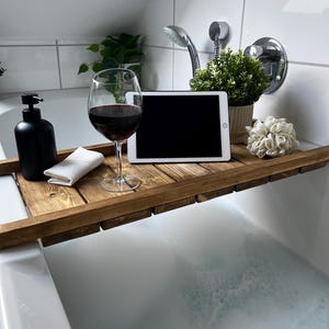 Puede incluir: Una bandeja de baño de madera sostiene una copa de vino tinto, un dispensador de jabón negro, una tableta blanca, una pequeña planta en maceta y un paño blanco. La bandeja abarca el ancho de una bañera blanca llena de burbujas.