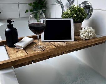 Puede incluir: Una bandeja de baño de madera sostiene una copa de vino tinto, un dispensador de jabón negro, una tableta blanca, una pequeña planta en maceta y un paño blanco. La bandeja abarca el ancho de una bañera blanca llena de burbujas.
