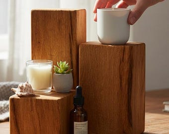May include: A display of home decor items on wooden blocks. Includes a small candle in a glass, a succulent in a pot, a brown bottle with a dropper, and a white ceramic cup. The blocks have a natural wood grain.