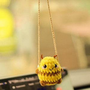 May include: A yellow and brown crocheted bee hanging from a rearview mirror in a car. The bee has a smiling face and black eyes.