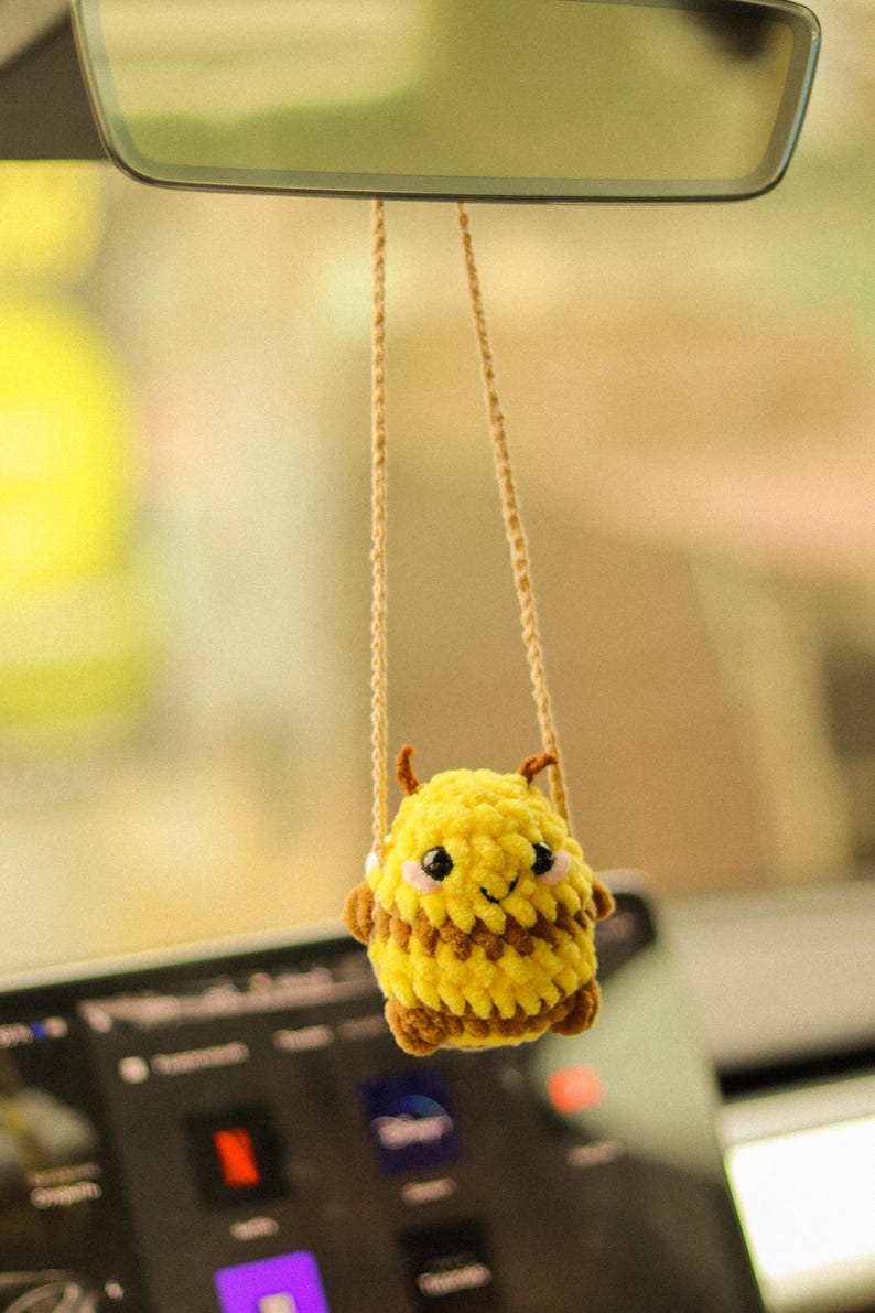 May include: A yellow and brown crocheted bee hanging from a rearview mirror in a car. The bee has a smiling face and black eyes.