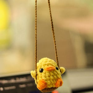 May include: A yellow crocheted chick hanging from a rearview mirror in a car. The chick has orange feet and a small orange beak.