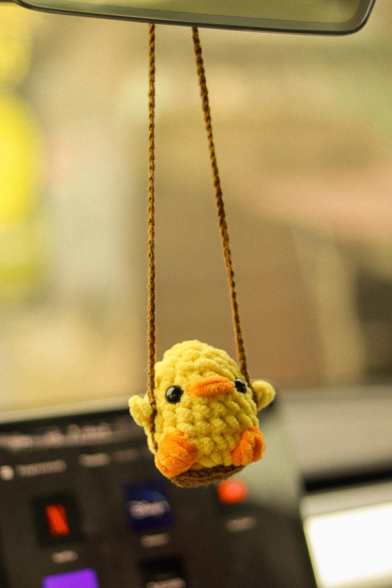 May include: A yellow crocheted chick hanging from a rearview mirror in a car. The chick has orange feet and a small orange beak.
