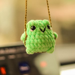May include: A green crocheted frog hanging from a brown string. The frog has black eyes and a small smile.