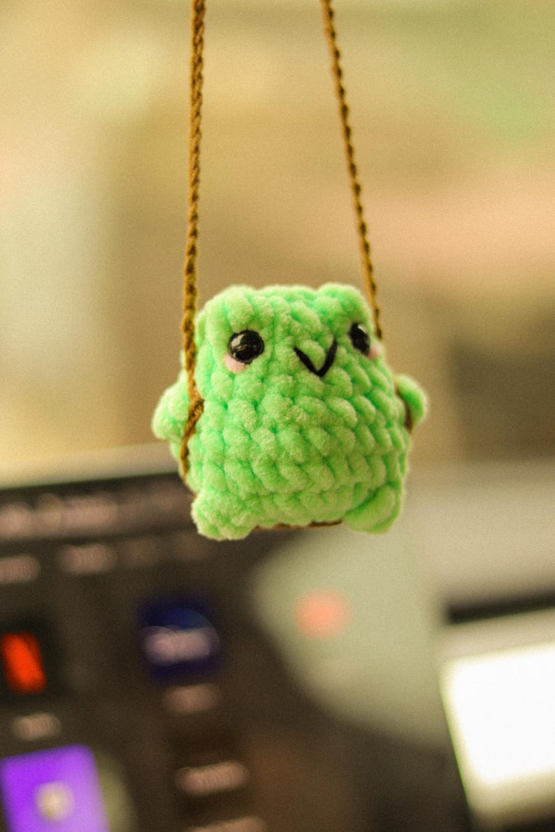 May include: A green crocheted frog hanging from a brown string. The frog has black eyes and a small smile.