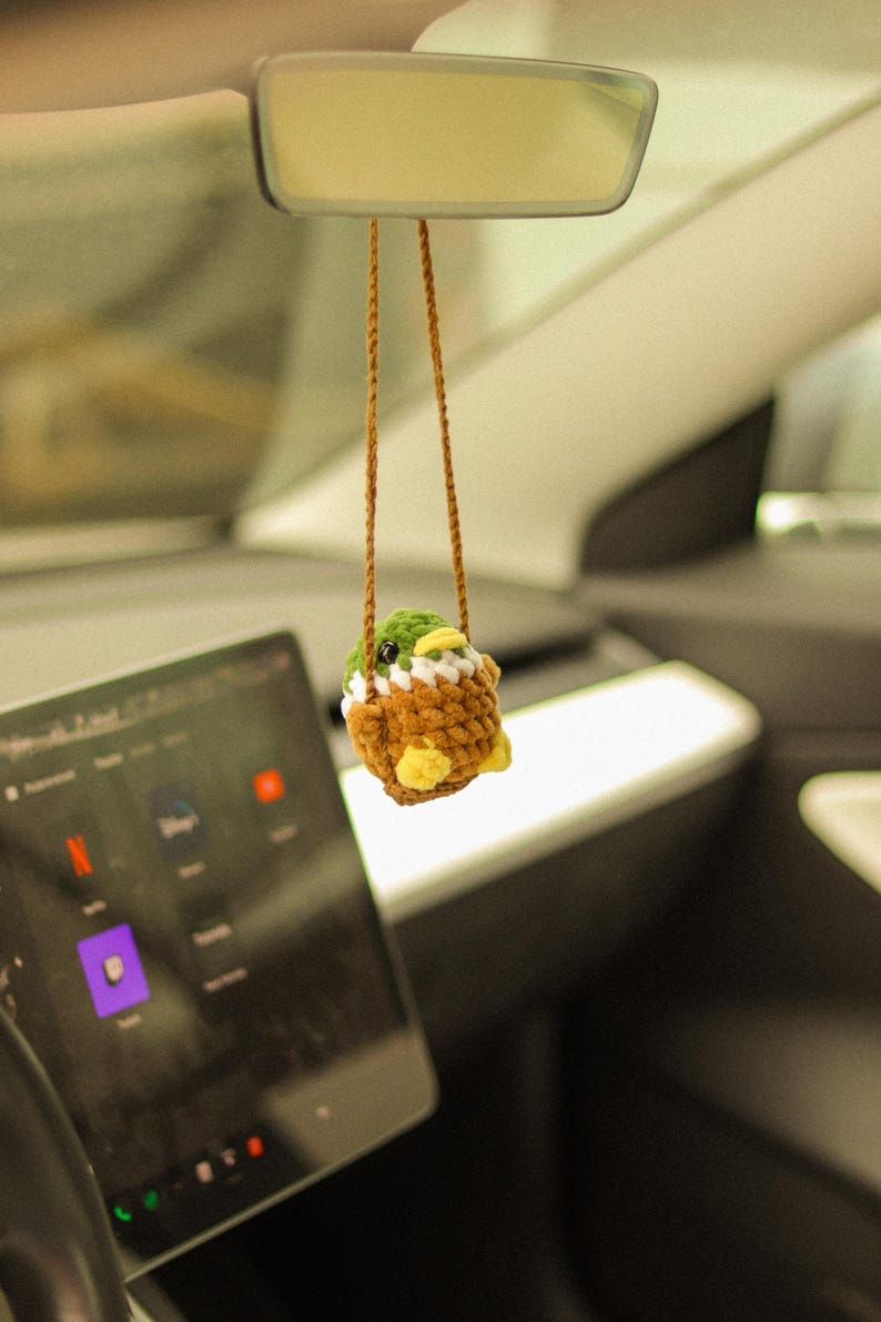 May include: A crocheted duck plush toy hanging from the rearview mirror of a car. The duck is brown, green, and white with yellow accents.