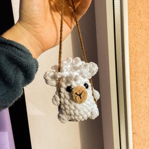 May include: A white crocheted sheep hanging from a brown string. The sheep has black eyes and a brown nose.