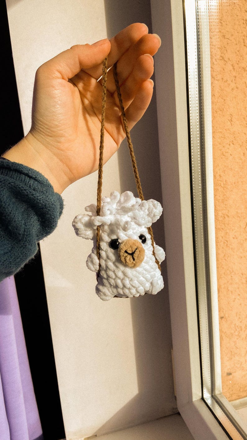 May include: A white crocheted sheep hanging from a brown string. The sheep has black eyes and a brown nose.