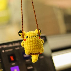 May include: A yellow crocheted plush toy of a cartoon character hanging from a brown string. The toy has a smiling face with red cheeks and is sitting on a swing.