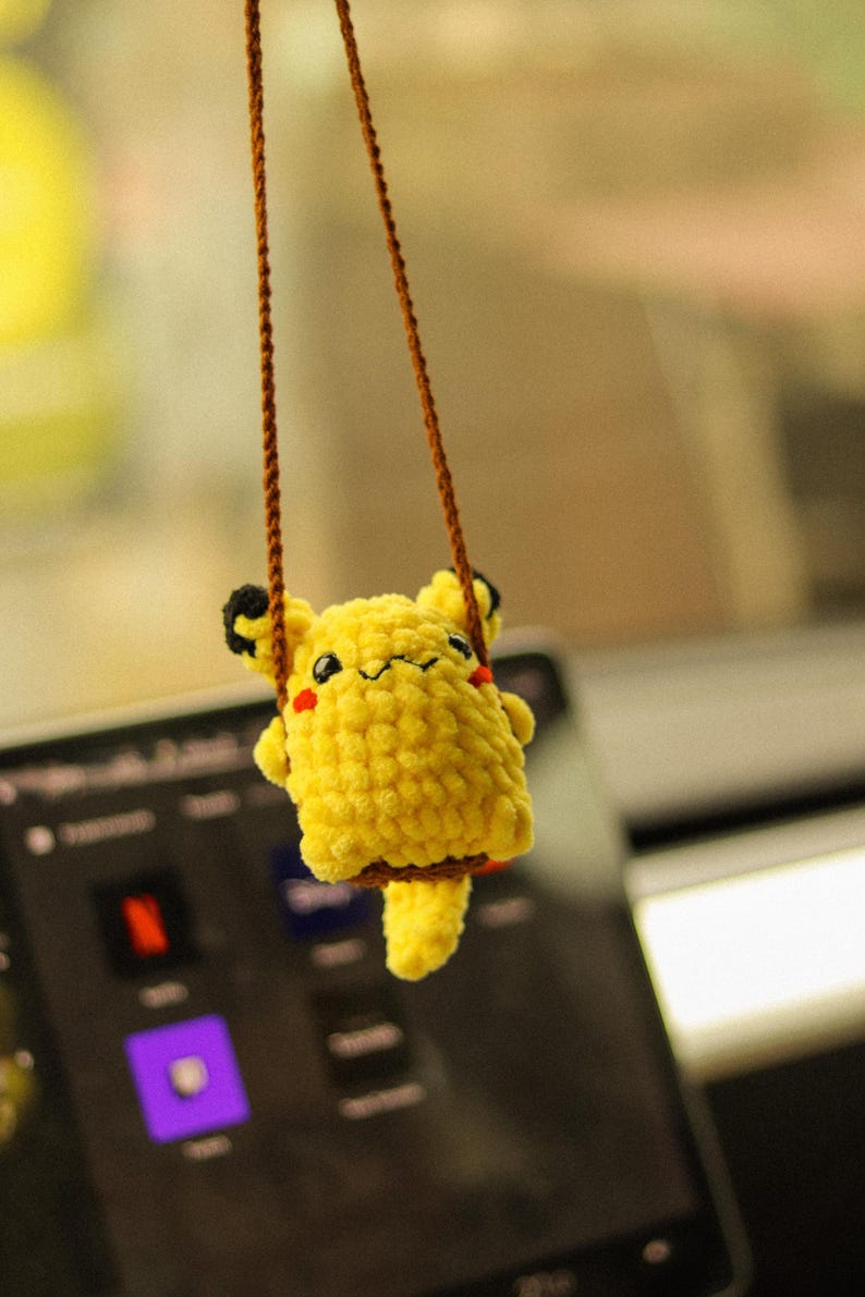 May include: A yellow crocheted plush toy of a cartoon character hanging from a brown string. The toy has a smiling face with red cheeks and is sitting on a swing.