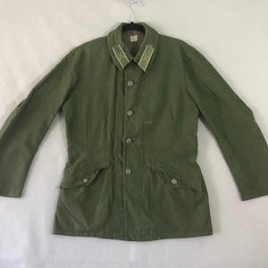 以下が含まれることがあります： ボタン留めのオリーブグリーンのジャケット。フロントに2つのポケットがあり、襟にはライトグリーンとイエローのアクセントが付いています。 ジャケットは金属製のハンガーに吊るされています。 ジャケットはヴィンテージのようです。