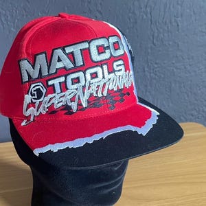 K&ouml;nnte beinhalten: Rote Baseballkappe mit dem Schriftzug "MATCO TOOLS SUPER NATIONALS" in silbernen und wei&szlig;en Buchstaben. Der Schirm ist schwarz mit wei&szlig;er und silberner Zierleiste. Die M&uuml;tze wird auf einem schwarzen Schaufensterkopf pr&auml;sentiert.