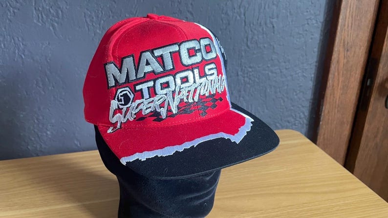 K&ouml;nnte beinhalten: Rote Baseballkappe mit dem Schriftzug "MATCO TOOLS SUPER NATIONALS" in silbernen und wei&szlig;en Buchstaben. Der Schirm ist schwarz mit wei&szlig;er und silberner Zierleiste. Die M&uuml;tze wird auf einem schwarzen Schaufensterkopf pr&auml;sentiert.