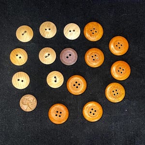 May include: A collection of fourteen circular wooden buttons in various shades of brown, ranging from light tan to dark mahogany. The buttons feature either two or four holes for attachment. A single copper-colored penny is included for scale.