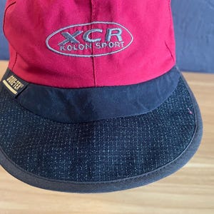 May include: A red and black baseball cap with the text "XCR KOLON SPORT" embroidered on the front. The cap has a curved brim and a "GORE-TEX" label.