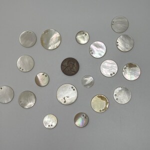 May include: A collection of seventeen circular mother-of-pearl buttons of varying sizes, each featuring two small holes for attachment. A single copper-colored United States penny is positioned near the center for scale, surrounded by the iridescent shell discs.