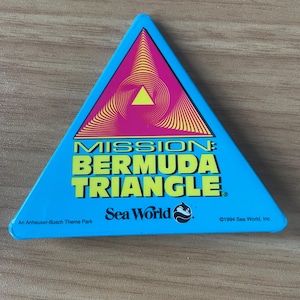 May include: A blue triangular souvenir from Sea World featuring the text "MISSION: BERMUDA TRIANGLE" in yellow. The design includes a pink and yellow graphic of a triangle within a triangle. The Sea World logo is also visible.