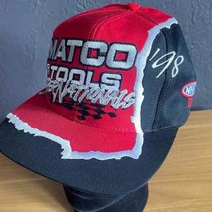 K&ouml;nnte beinhalten: Rote, schwarze und wei&szlig;e Baseballkappe mit dem Text "MATCO TOOLS INTERNATIONALS" und dem Jahr "'98". Die Kappe hat einen gebogenen Schirm und eine strukturierte Krone mit einem karierten Flaggenmuster.