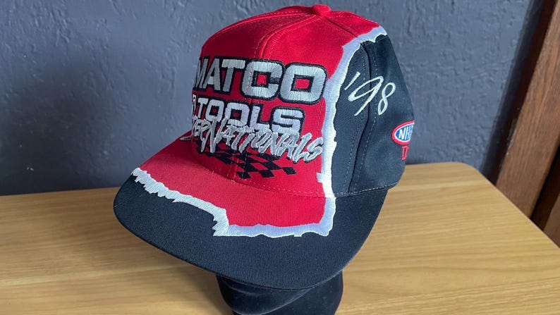 K&ouml;nnte beinhalten: Rote, schwarze und wei&szlig;e Baseballkappe mit dem Text "MATCO TOOLS INTERNATIONALS" und dem Jahr "'98". Die Kappe hat einen gebogenen Schirm und eine strukturierte Krone mit einem karierten Flaggenmuster.