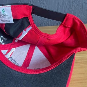 K&ouml;nnte beinhalten: Eine schwarze und rote Baseballkappe mit gebogenem Schirm. Die Innenseite hat ein wei&szlig;es und schwarzes Design und ein Etikett mit der Aufschrift "Kuztu". Die Kappe besteht aus einer Polyester- und Baumwollmischung.