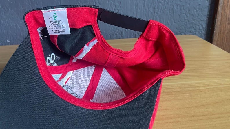 K&ouml;nnte beinhalten: Eine schwarze und rote Baseballkappe mit gebogenem Schirm. Die Innenseite hat ein wei&szlig;es und schwarzes Design und ein Etikett mit der Aufschrift "Kuztu". Die Kappe besteht aus einer Polyester- und Baumwollmischung.