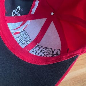 K&ouml;nnte beinhalten: Eine rot-schwarze Baseballkappe mit wei&szlig;em Text im Inneren. Die Kappe hat einen schwarzen Schirm und ein rotes Innenfutter. Im Inneren befinden sich wei&szlig;er Text und ein wei&szlig;es Mesh-Futter. Die Kappe ist aus strapazierf&auml;higem Stoff gefertigt.