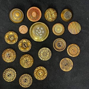 May include: A collection of sixteen round metal buttons in various shades of gold, bronze, and copper, featuring intricate embossed patterns, floral motifs, and geometric textures, displayed alongside a single copper-colored United States penny.