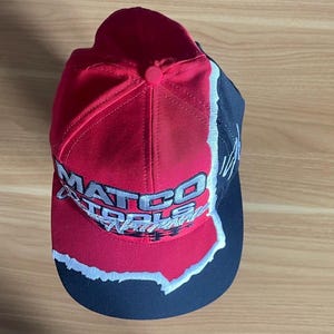 K&ouml;nnte beinhalten: Eine rot, schwarz und wei&szlig;e Baseballkappe mit dem Schriftzug "MATCO TOOLS" in wei&szlig;en Buchstaben. Die Kappe hat einen gebogenen Schirm und einen Knopf oben. Das Design zeigt einen zerrissenen Papiereffekt.