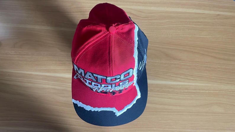 K&ouml;nnte beinhalten: Eine rot, schwarz und wei&szlig;e Baseballkappe mit dem Schriftzug "MATCO TOOLS" in wei&szlig;en Buchstaben. Die Kappe hat einen gebogenen Schirm und einen Knopf oben. Das Design zeigt einen zerrissenen Papiereffekt.