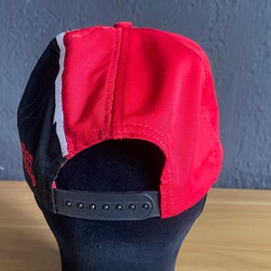 K&ouml;nnte beinhalten: Rote und schwarze Baseballkappe mit wei&szlig;em Streifen. Die Kappe hat einen schwarzen, verstellbaren Riemen und einen gebogenen Schirm. Die R&uuml;ckseite der Kappe ist rot, die Seiten sind schwarz mit einem wei&szlig;en Streifen.